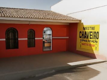 Fachada da loja Mestre Chaveiro, com parede vermelha, telhado de telhas e um grande letreiro amarelo na lateral com o texto 'MESTRE CHAVEIRO, CÓPIAS/AFIAÇÃO, 99777-0750'.