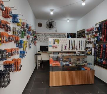 Visão interna da loja Mestre Chaveiro. Ao fundo, um grande painel branco com centenas de chaves virgens. No centro, um balcão de atendimento em vidro e madeira. Nas paredes, prateleiras com diversos produtos como chaveiros e fechaduras.